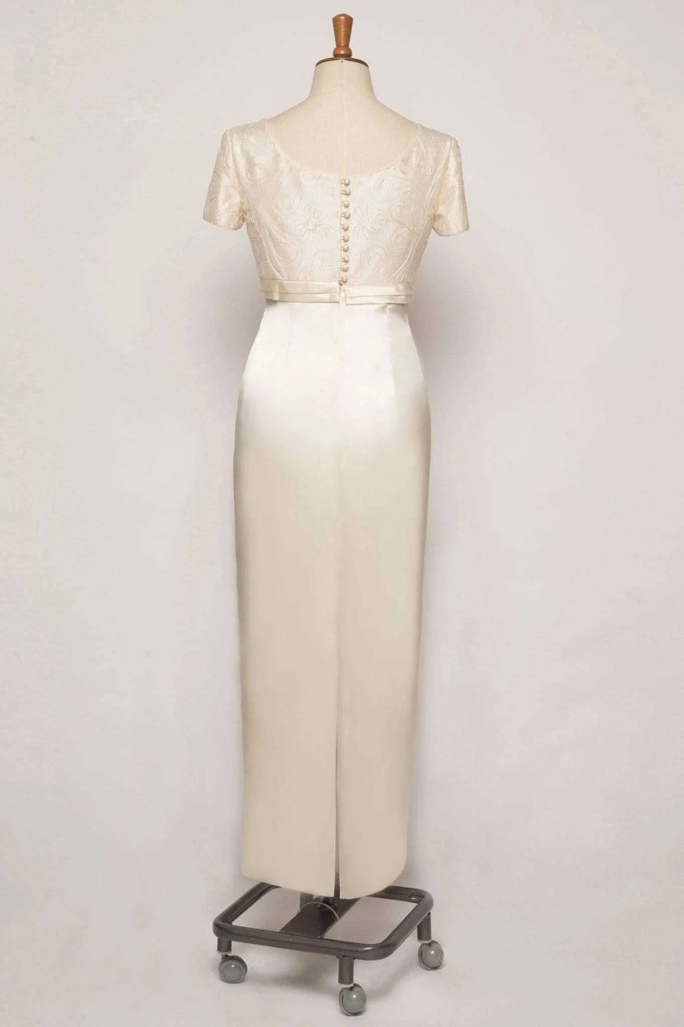 A sophisticated satin empire column-style dress featuring short sleeves, a square neckline, pleat detailing, piped edges, and covered buttons, crafted from embroidered pure silk and satin.