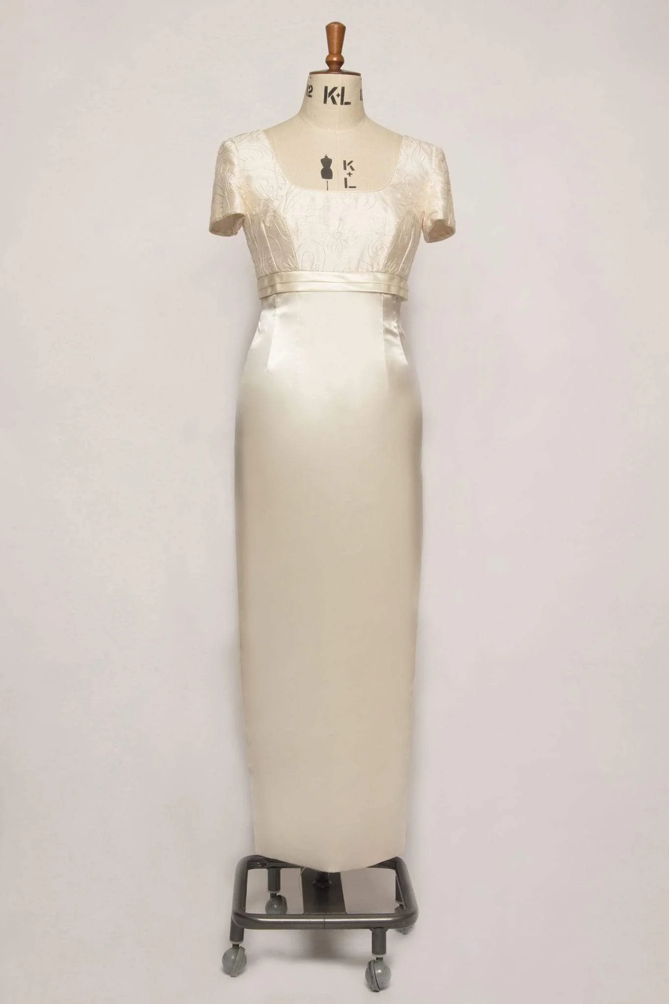 A sophisticated satin empire column-style dress featuring short sleeves, a square neckline, pleat detailing, piped edges, and covered buttons, crafted from embroidered pure silk and satin.