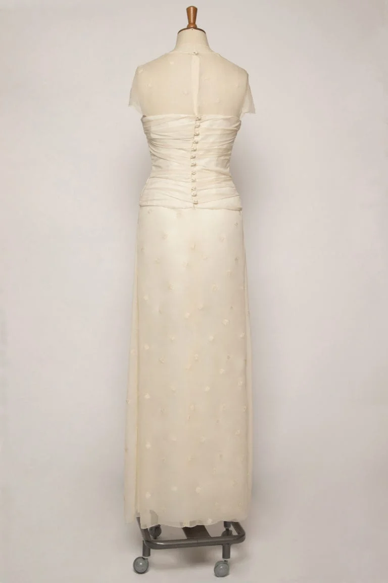 Back view of Long flowing A-line chiffon dress with ruching, a sheer overlay top, a dropped waist, and delicate embroidery. Showing covered button and loop fastening detailing at the back.